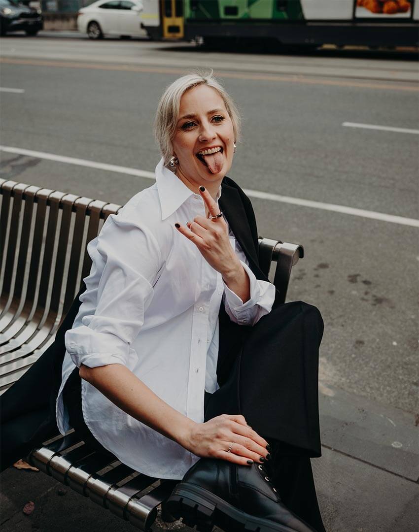 Business woman sat on a bench in Melbourne wearing a white shirt and sticking her tongue out.